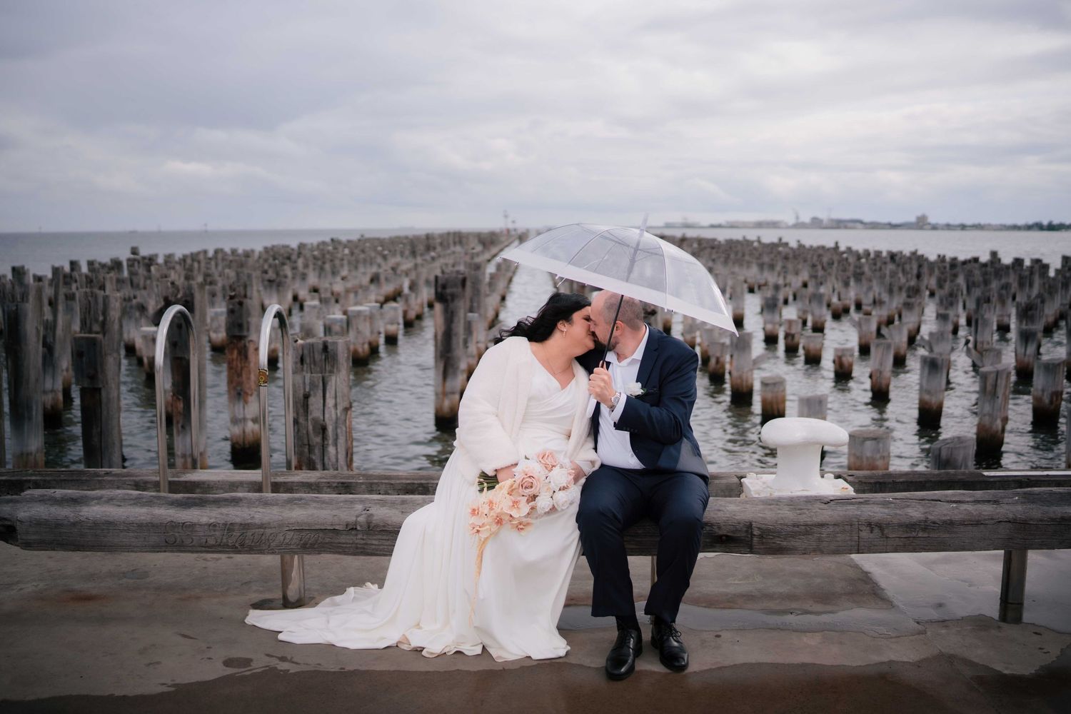 Bride and groom share a kiss under a clear umbrella while seated on a rainy waterfront pier.