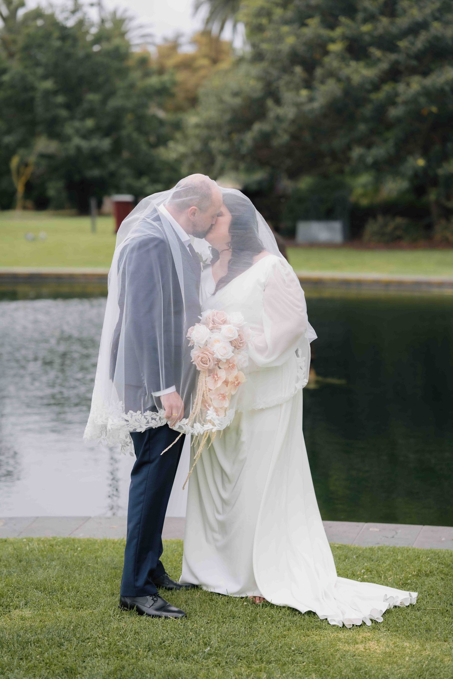Bride and groom kissing under a white veil by a pond in a lush green garden.