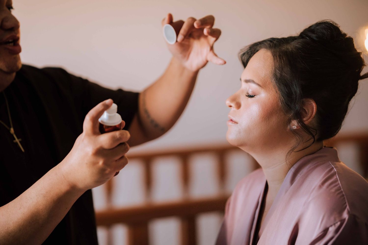 Makeup artist applying setting spray to bride wearing pink robe during wedding preparations.