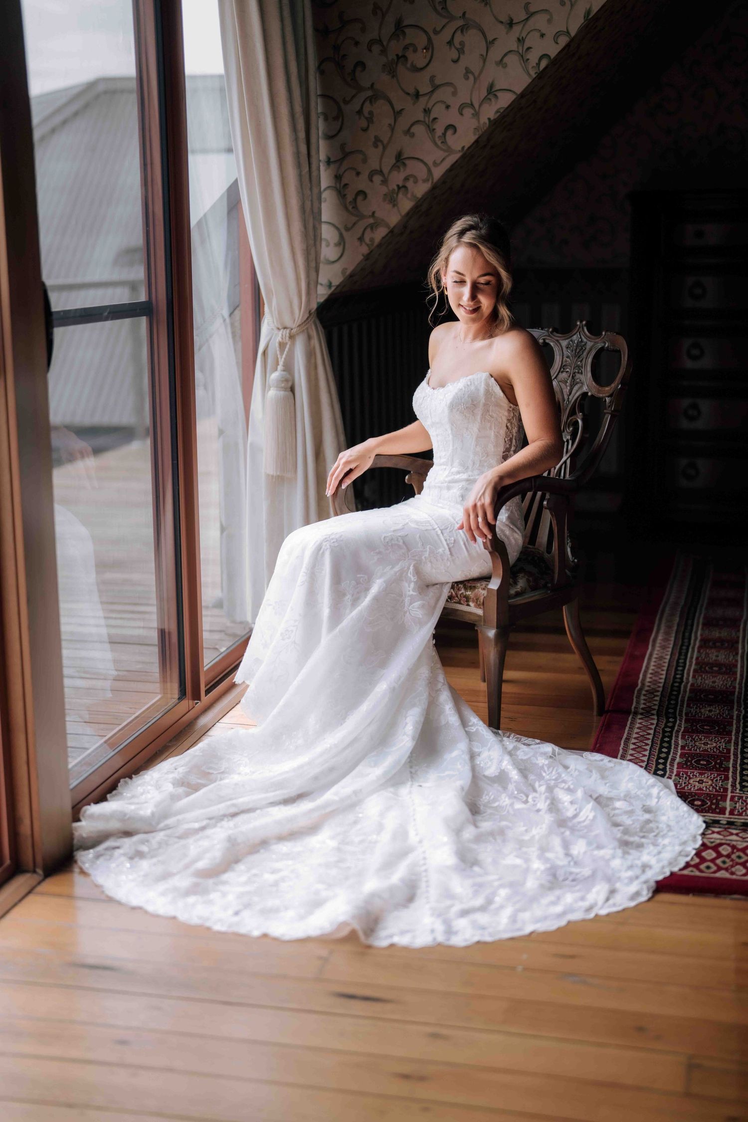 Bride in fitted lace wedding gown with train sits gracefully by large window in chair.