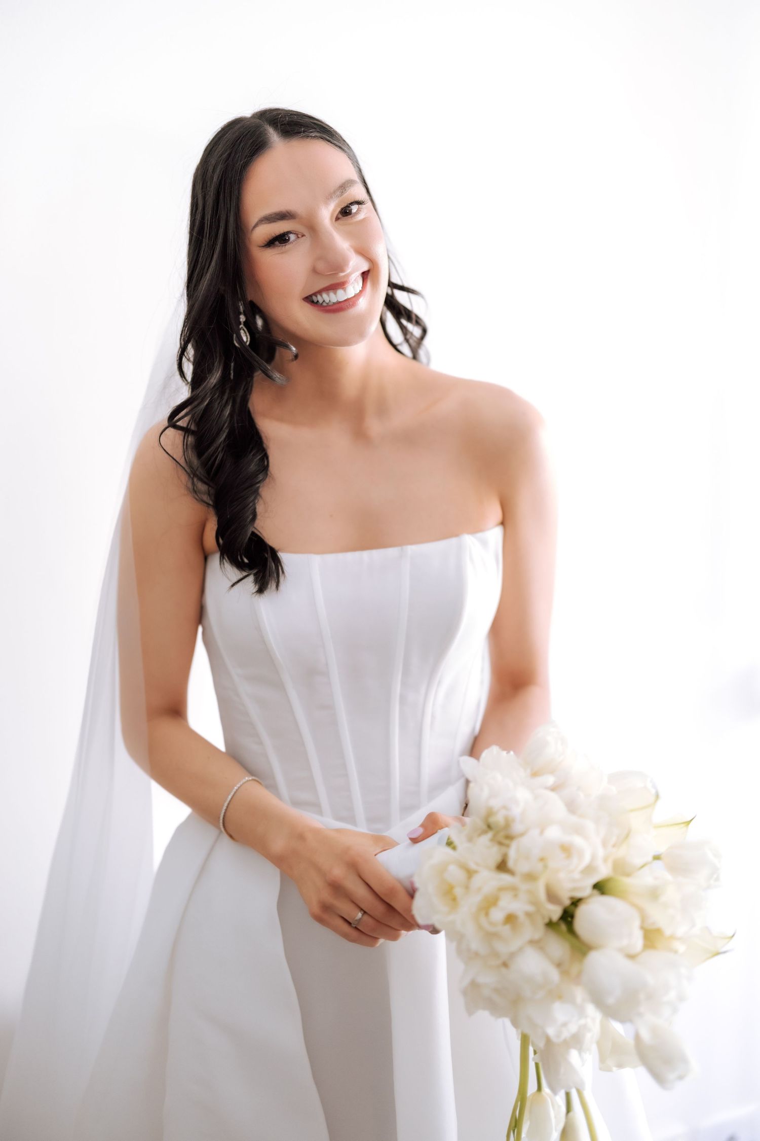 Smiling bride in a strapless white gown holding a bouquet of white tulips with a veil and elegant earrings.