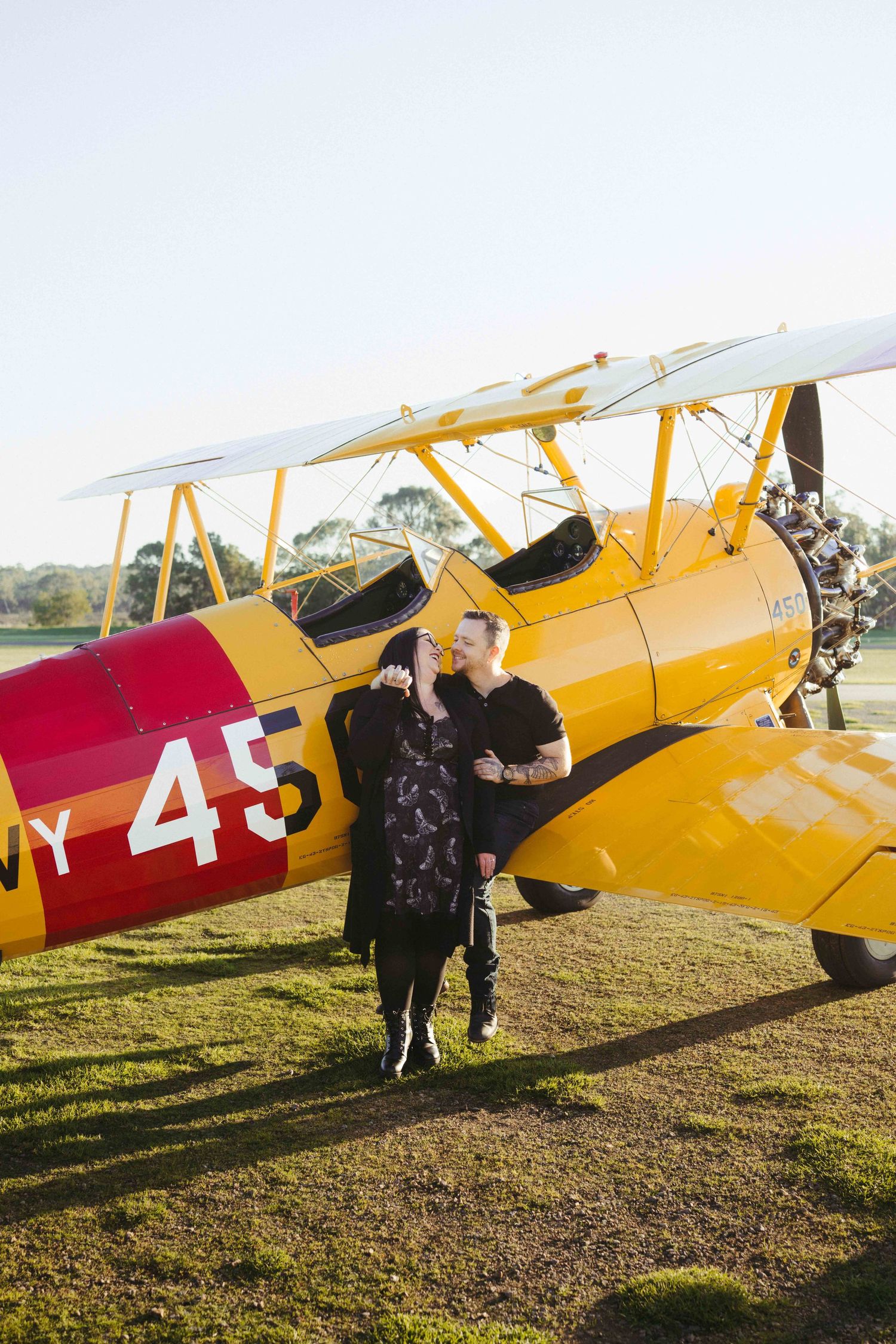 Couple embracing in front of a vintage yellow and red biplane numbered 45 on a sunny green airfield.