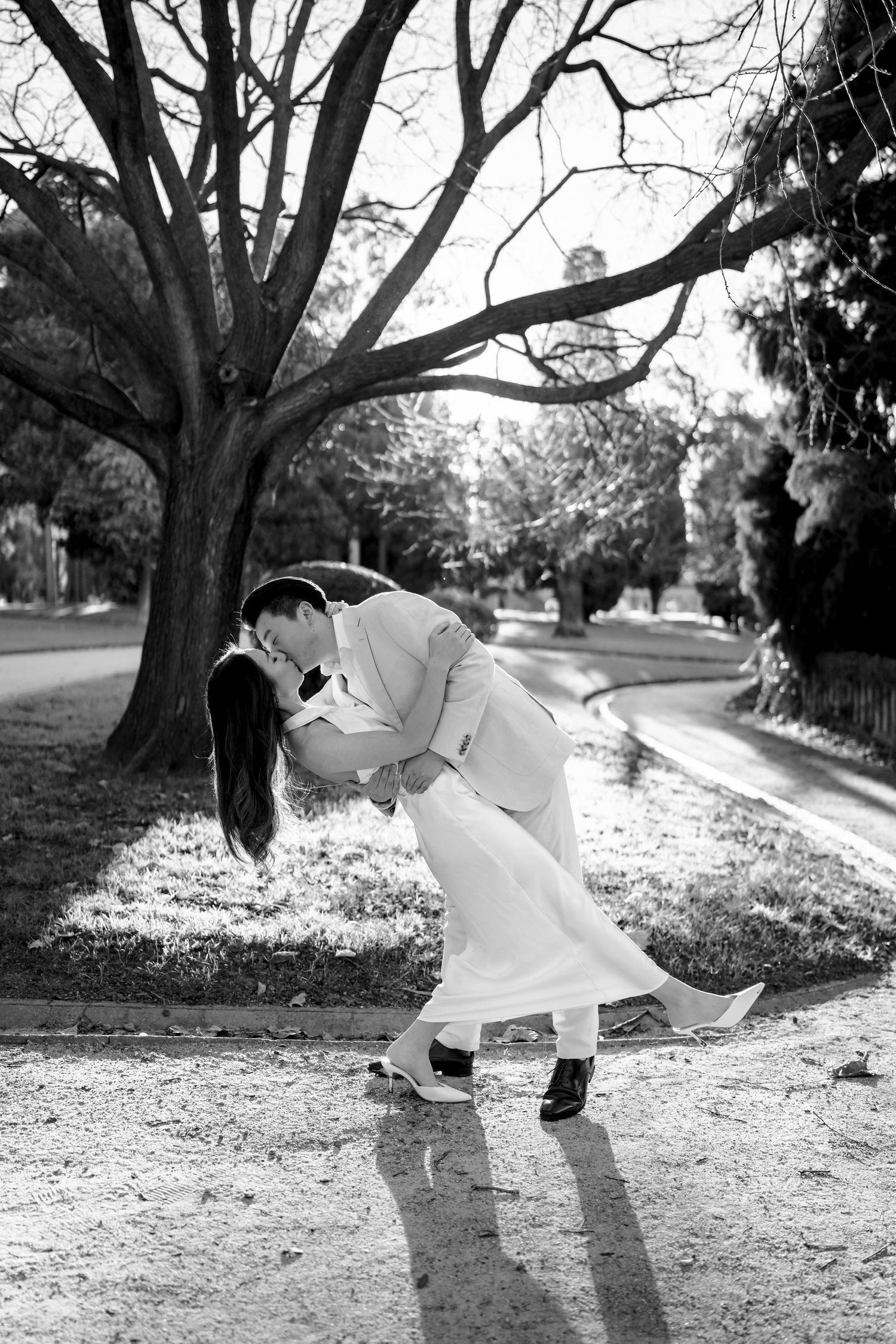 Black and white photo of a couple sharing a romantic dip kiss on a tree-lined path, bride in white dress.