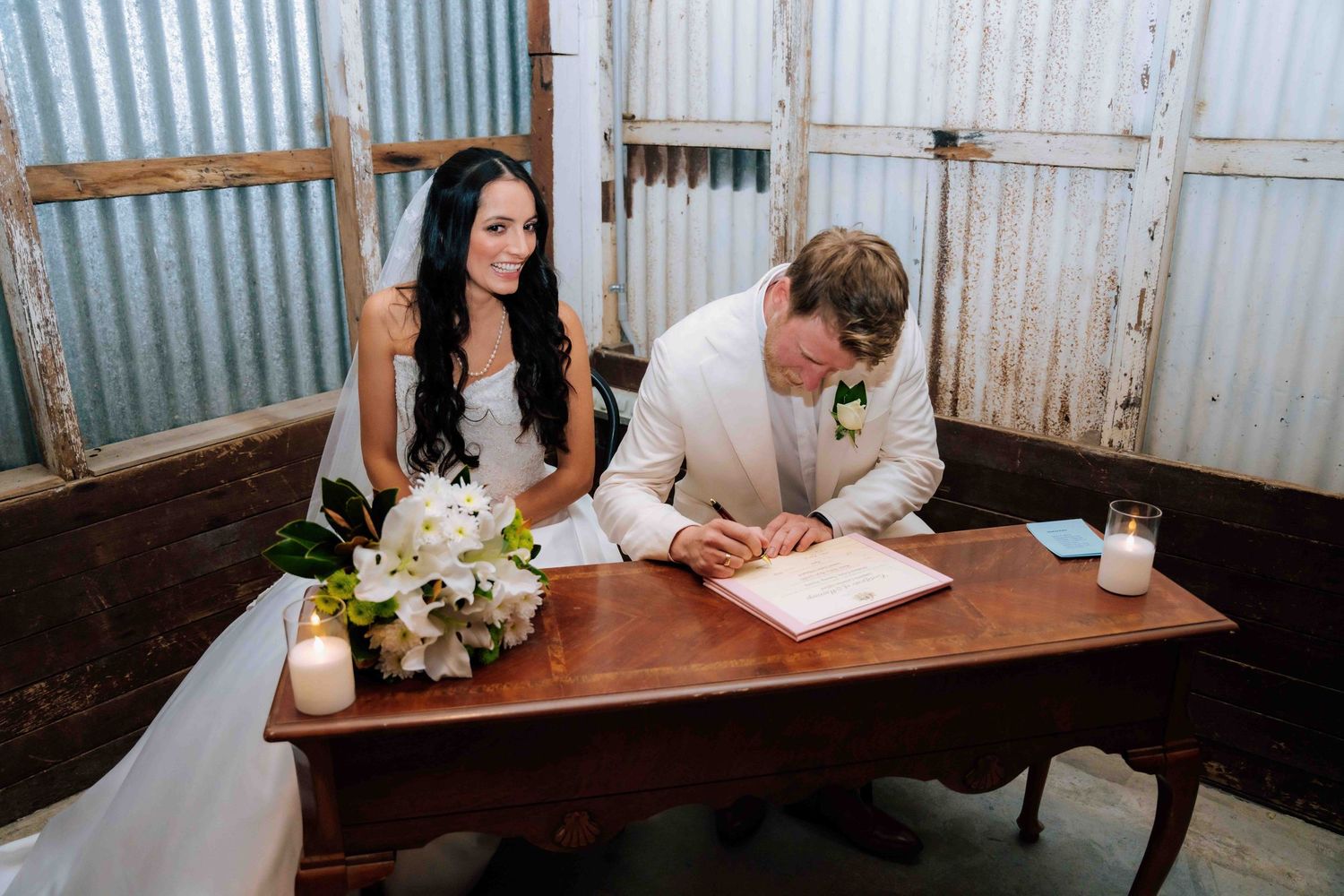 Bride and groom signing wedding registry at wooden desk in rustic barn venue with white floral bouquet and candles.