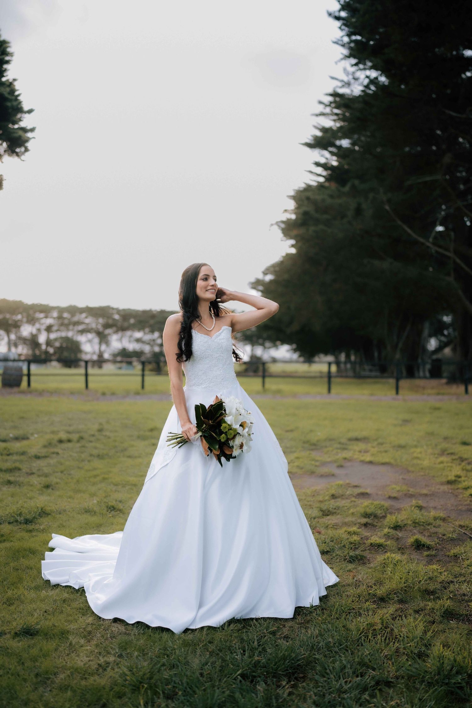 Bride in elegant white ball gown with train holds bouquet while posing alone in a green field at her wedding.