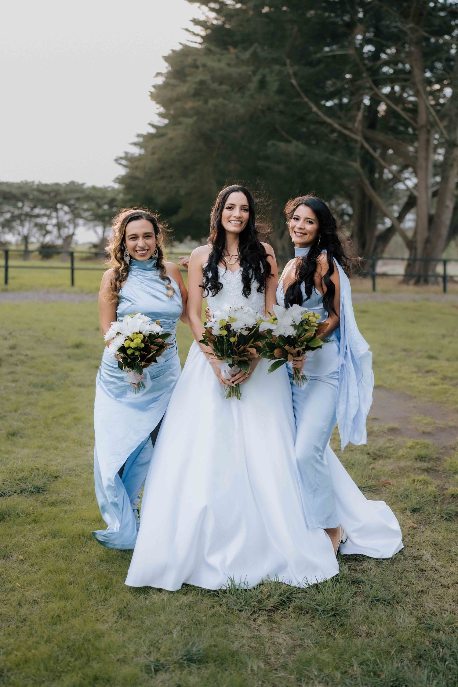 Bride in white gown poses with two bridesmaids in light blue dresses holding bouquets outdoors.