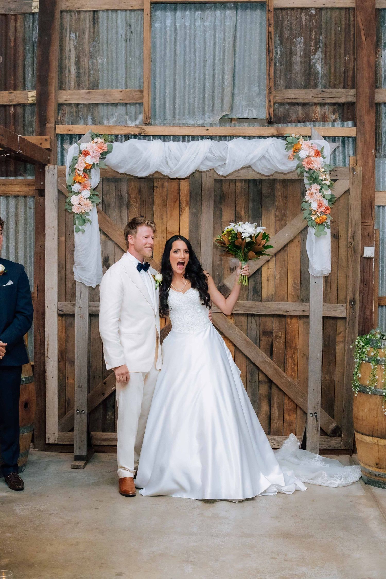 Bride and groom celebrating joyfully at rustic barn altar decorated with floral arrangements and draping.