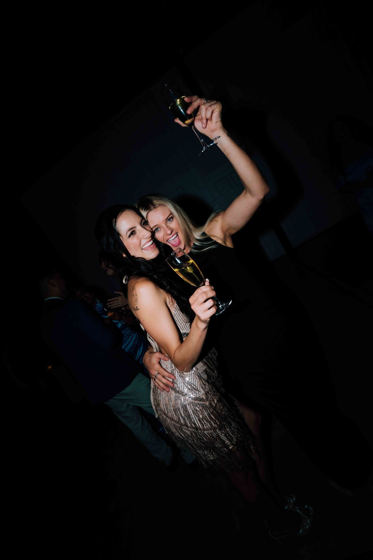 Two women celebrating at party holding champagne glasses in dark venue.