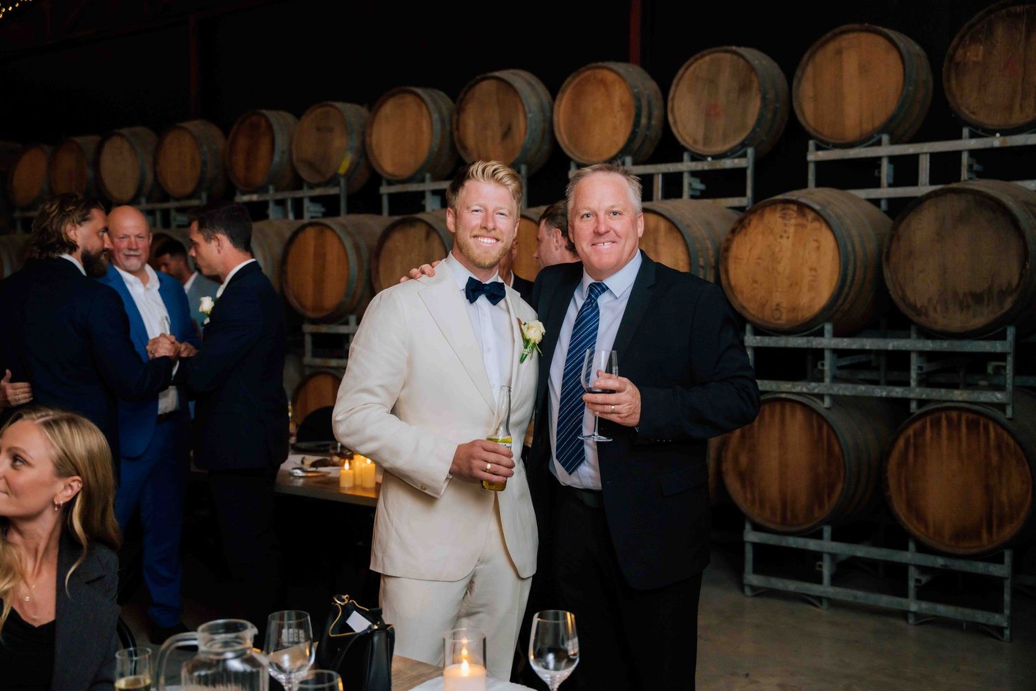 Two men in suits smile together at wedding reception beside oak wine barrels.
