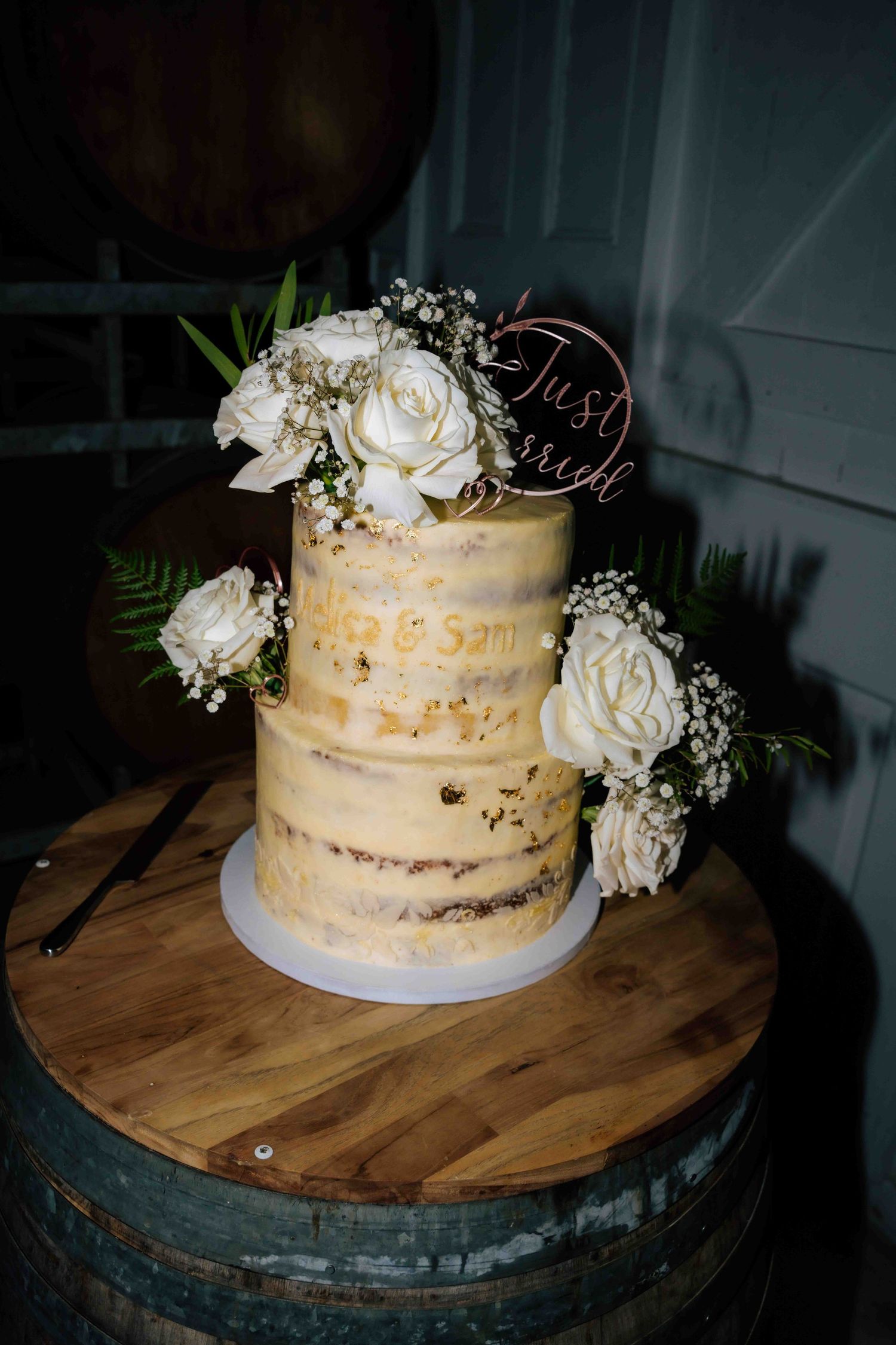 Two-tier semi-naked wedding cake decorated with white roses and a Just Married topper on a wooden barrel.