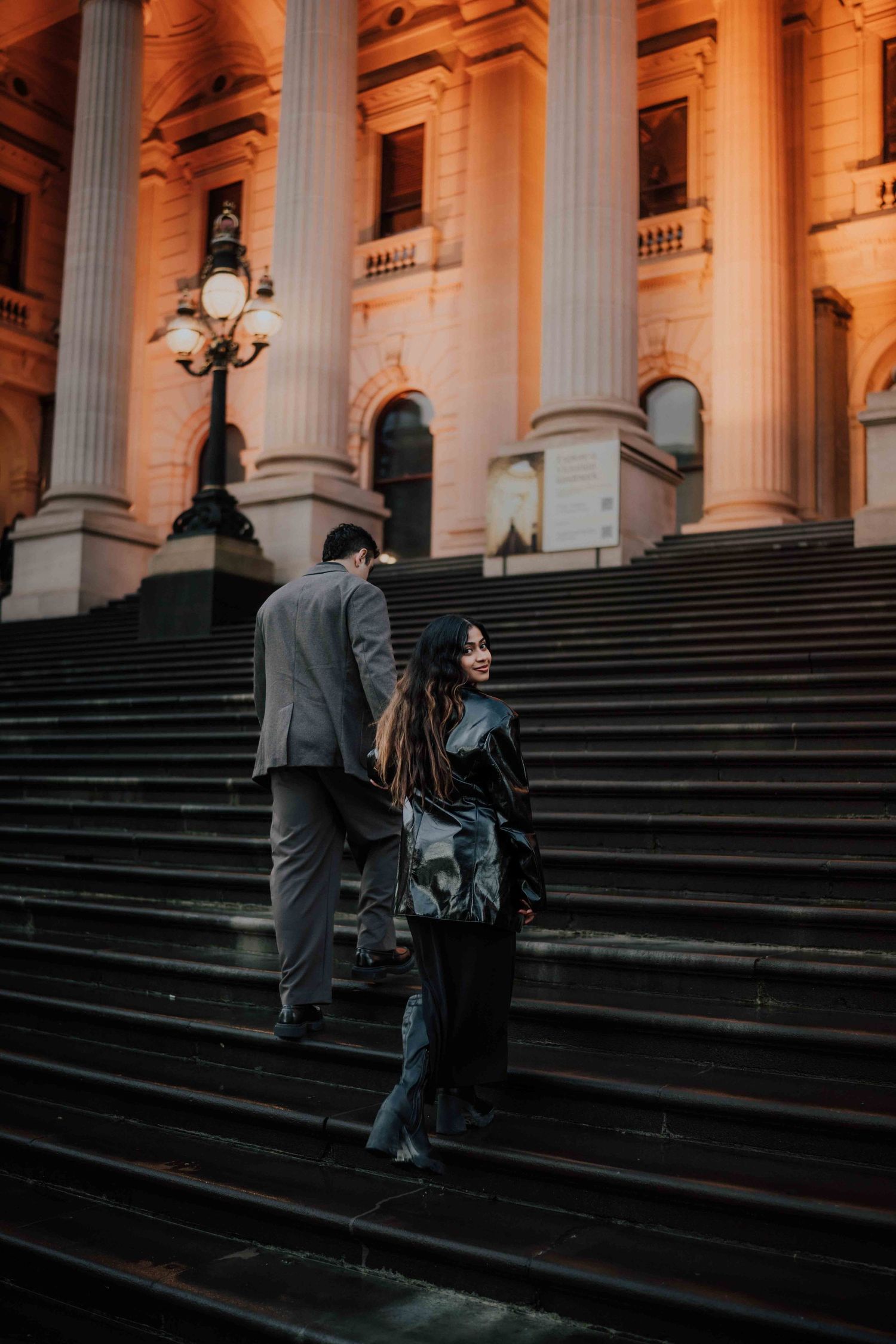 Woman in leather jacket looks back while man walks up steps of a grand columned building.
