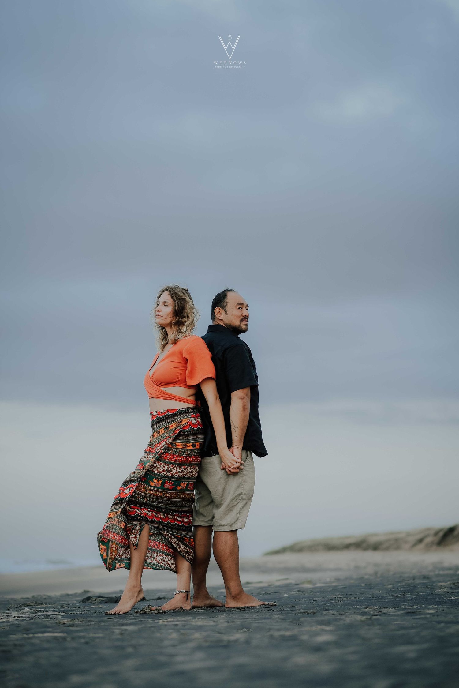 Couple standing back-to-back holding hands on a beach, woman in orange top and colorful skirt.