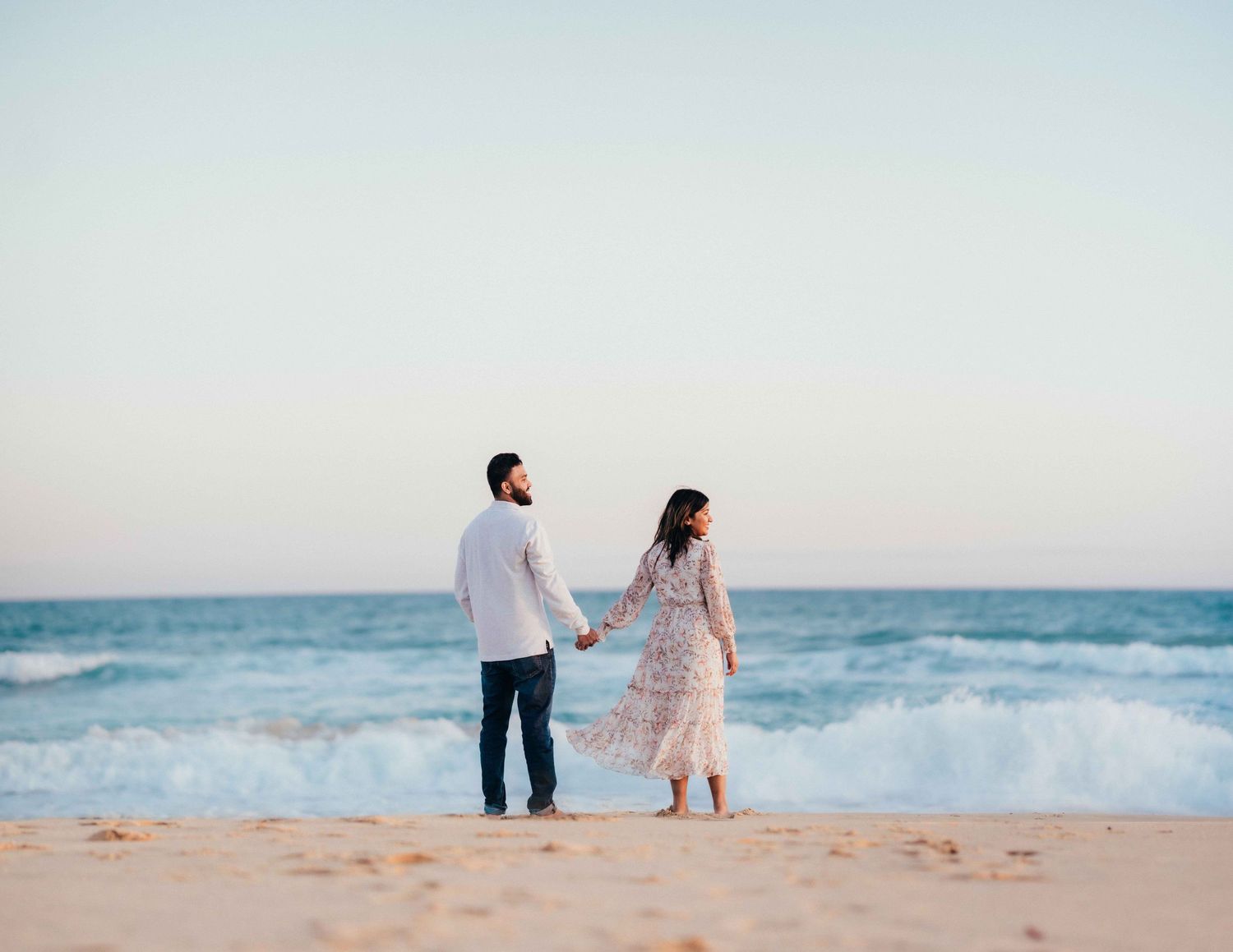 Couple holding hands walking toward ocean waves on a sandy beach at golden hour.