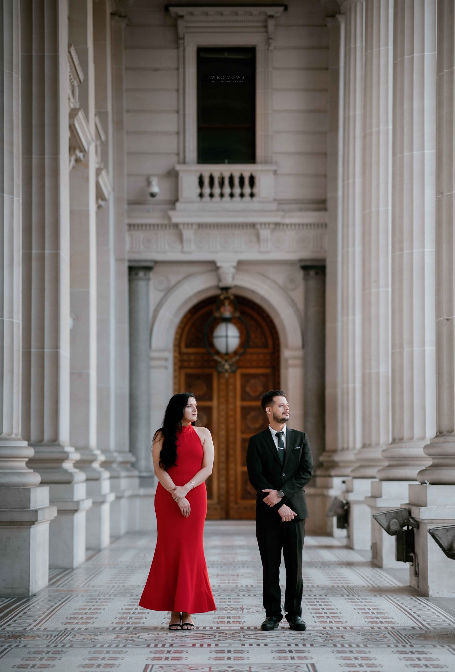 Elegant couple in red dress and black suit stand apart beneath grand columns and arched doorway.