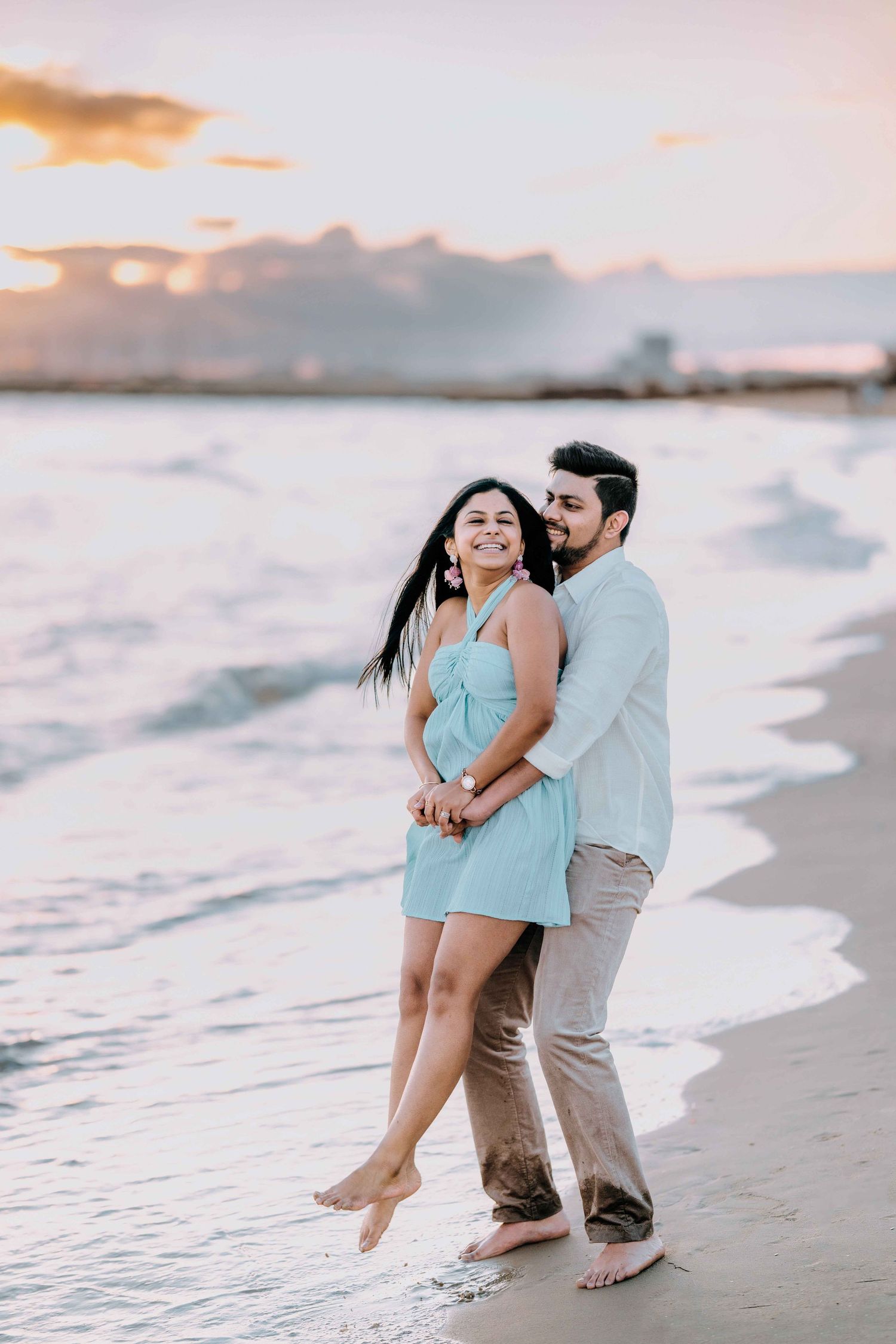 Happy couple embracing on a sandy beach at sunset, woman in turquoise dress smiling joyfully.