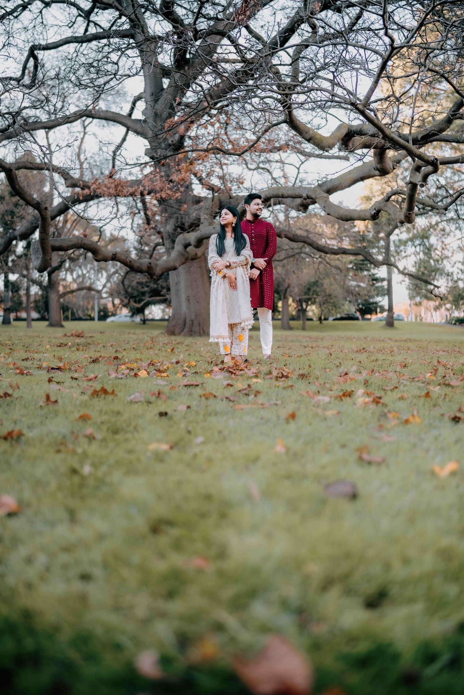 Couple in traditional Indian dress standing together under bare winter trees on a leaf-covered lawn.