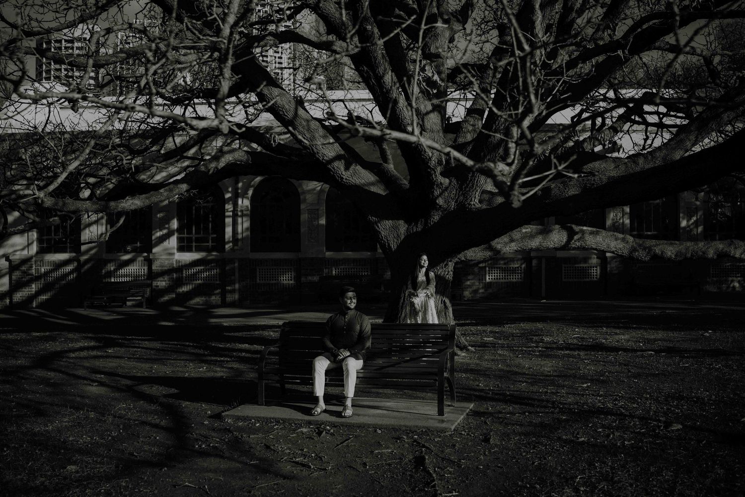 Black and white photo of couple sitting on bench beneath large bare tree in park.