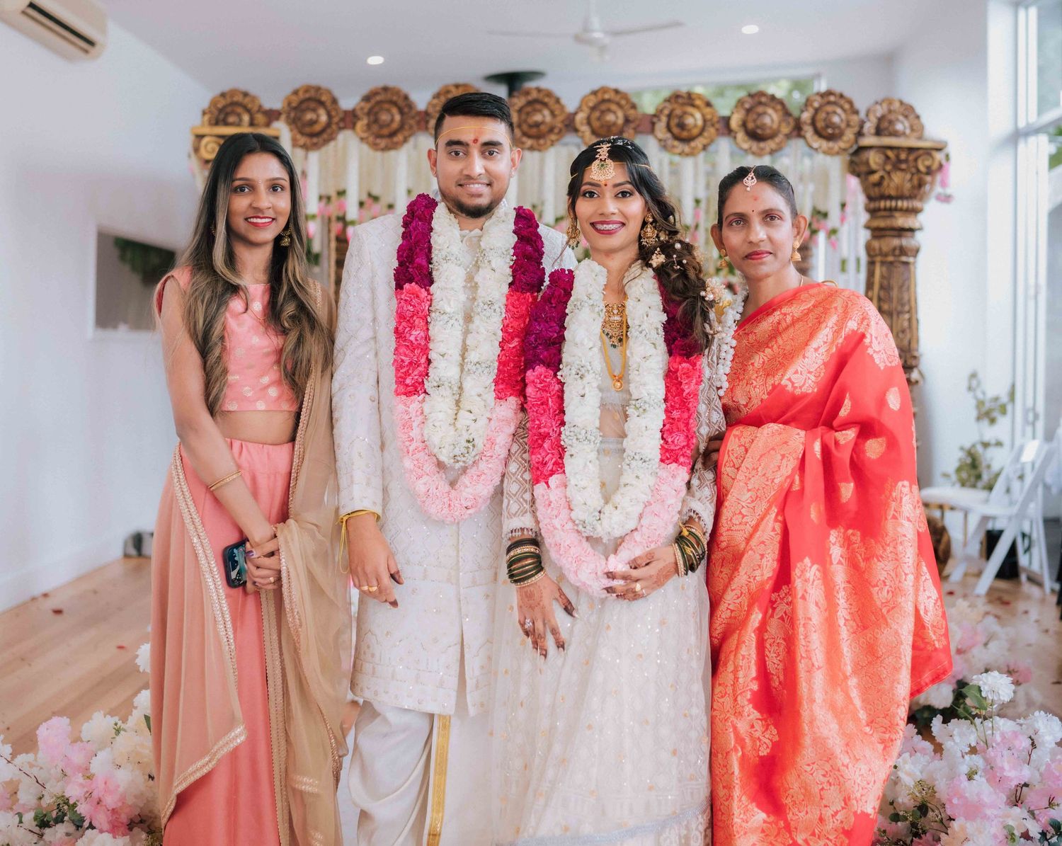 Indian bride and groom wearing floral garlands posing with two female relatives at a decorated wedding ceremony.