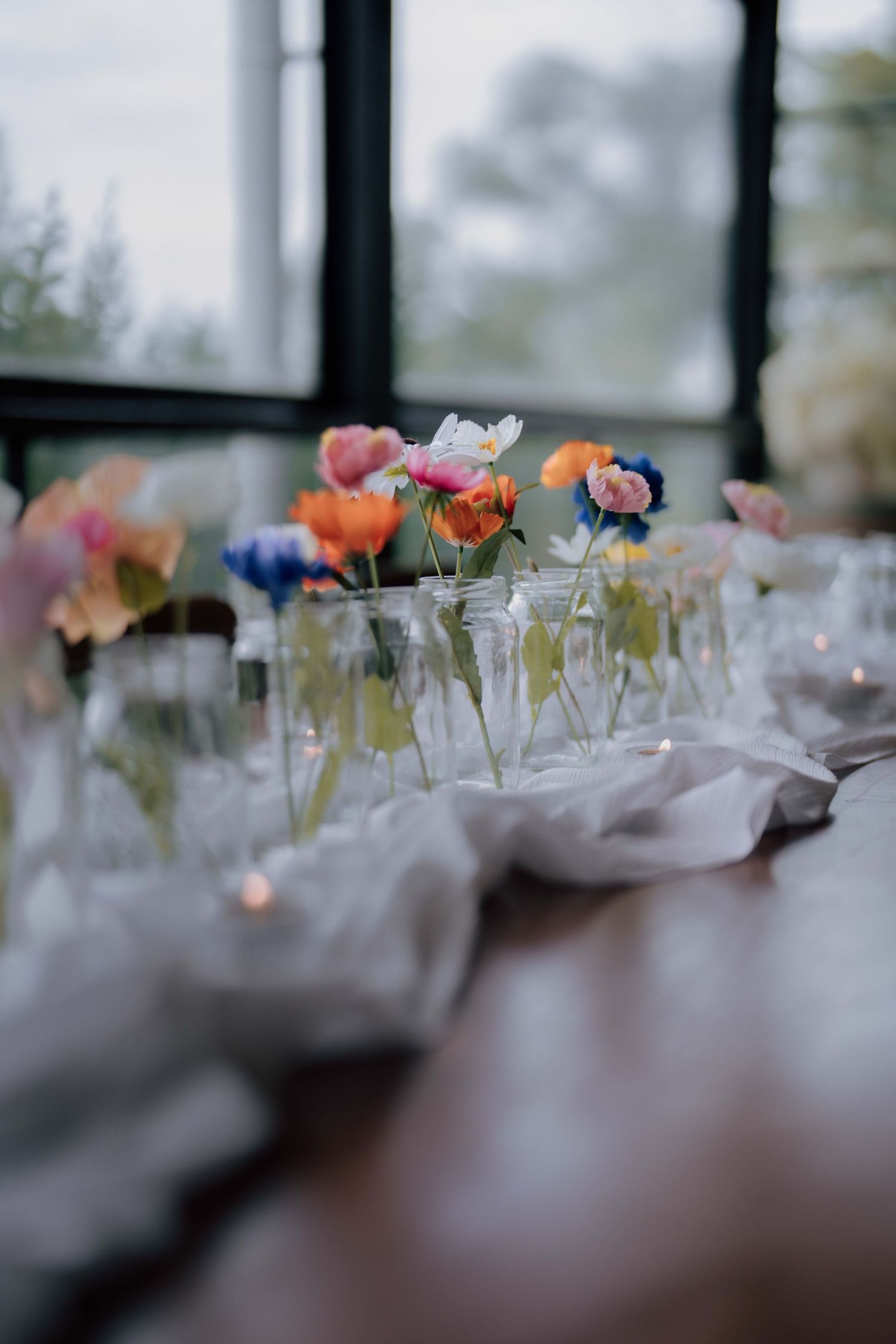 Colorful poppies and wildflowers in glass bud vases along a white linen runner on a wooden reception table.