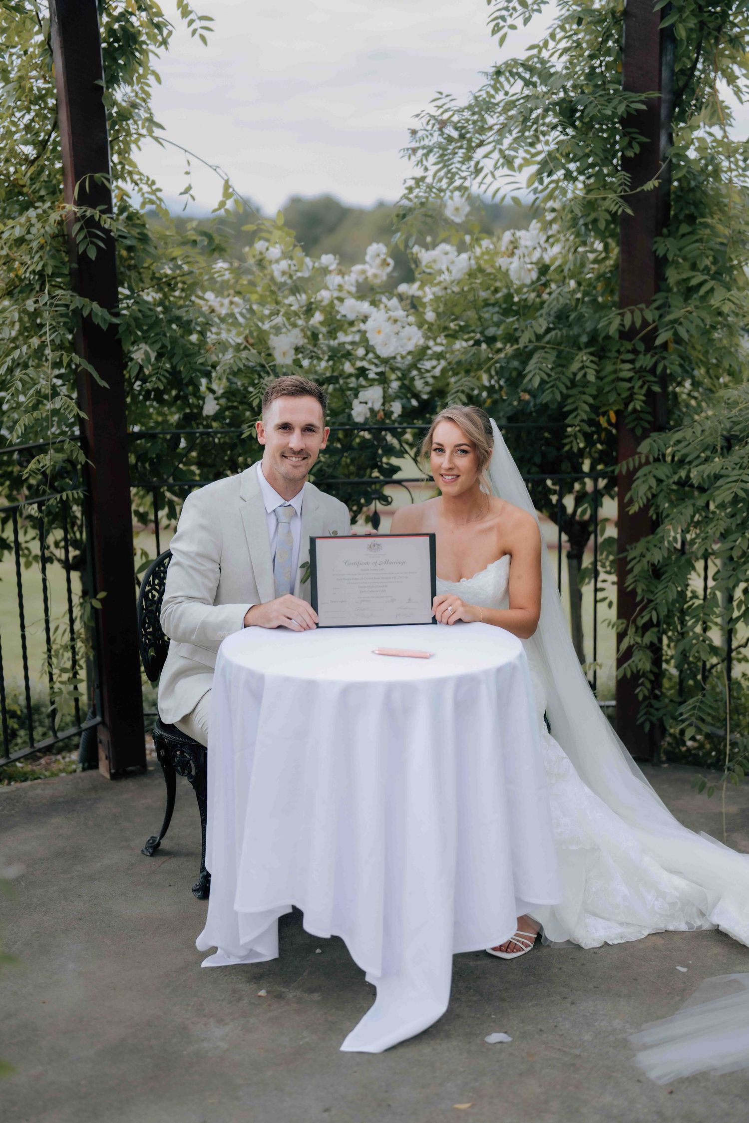 Bride and groom sitting at white table holding marriage certificate outdoors at garden wedding ceremony.
