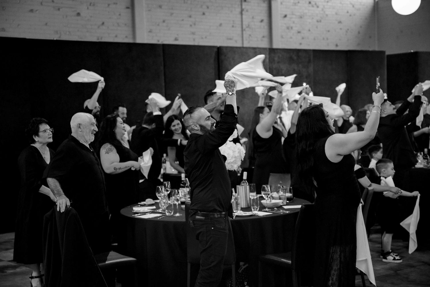 Black and white photo of wedding guests waving white napkins in celebration at reception dinner.