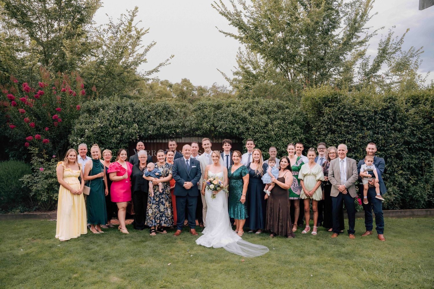 Large family group portrait at wedding with bride and groom centered on green lawn with flowering trees behind.