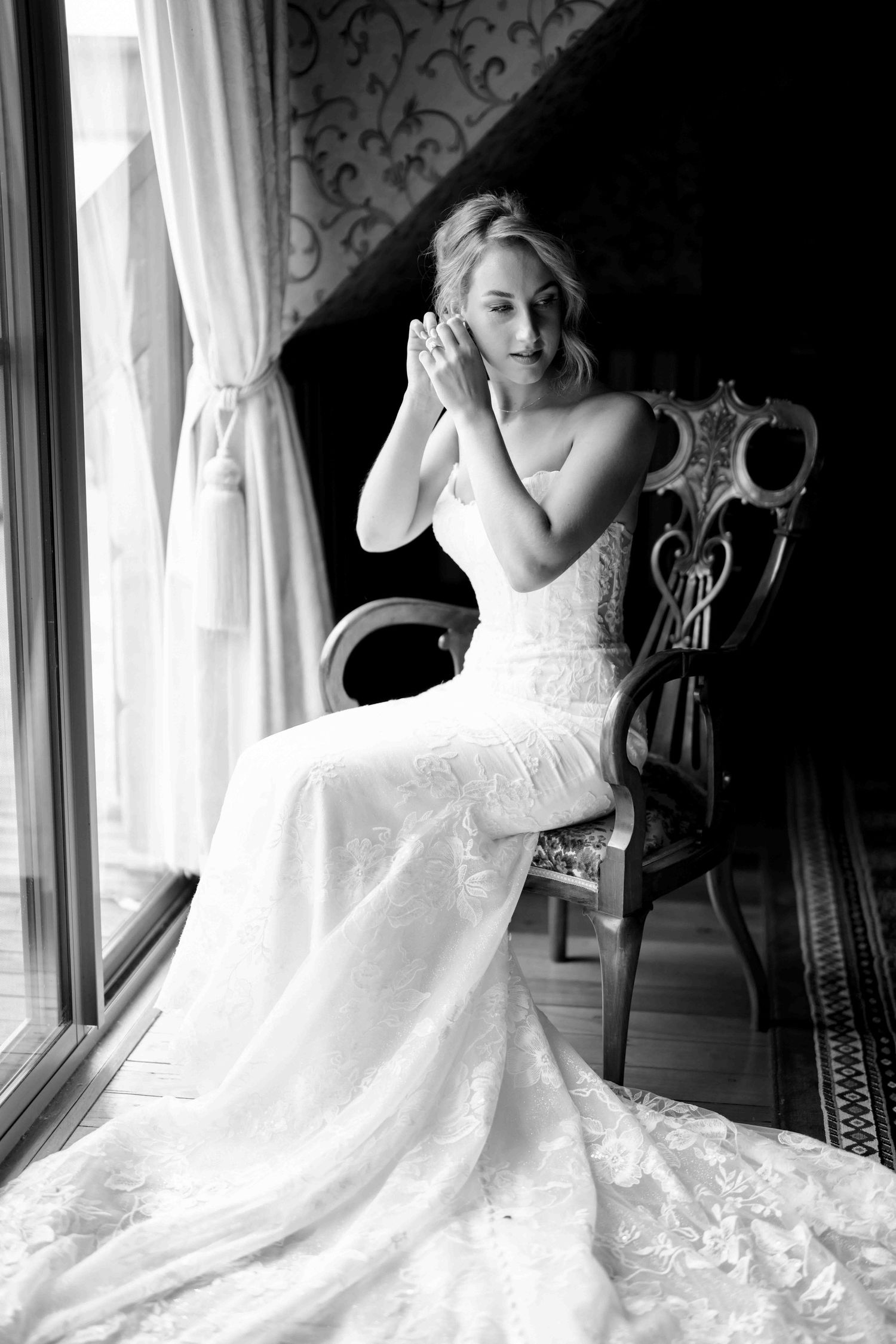 Black and white portrait of a bride in a lace gown adjusting her earring while seated in an antique chair.
