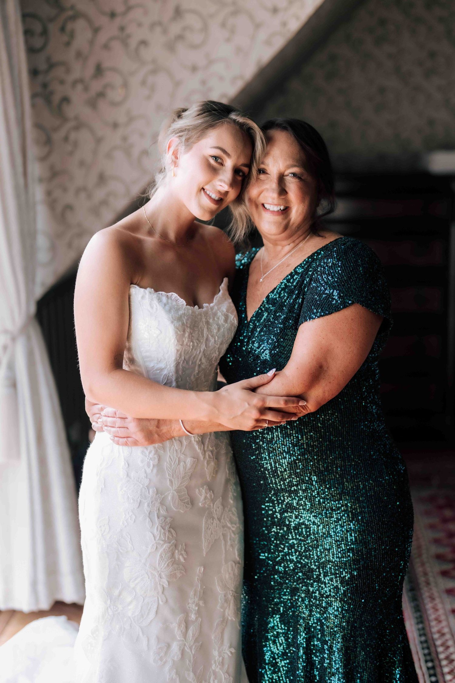 Bride in lace wedding gown posing with smiling mother in teal sequin dress.