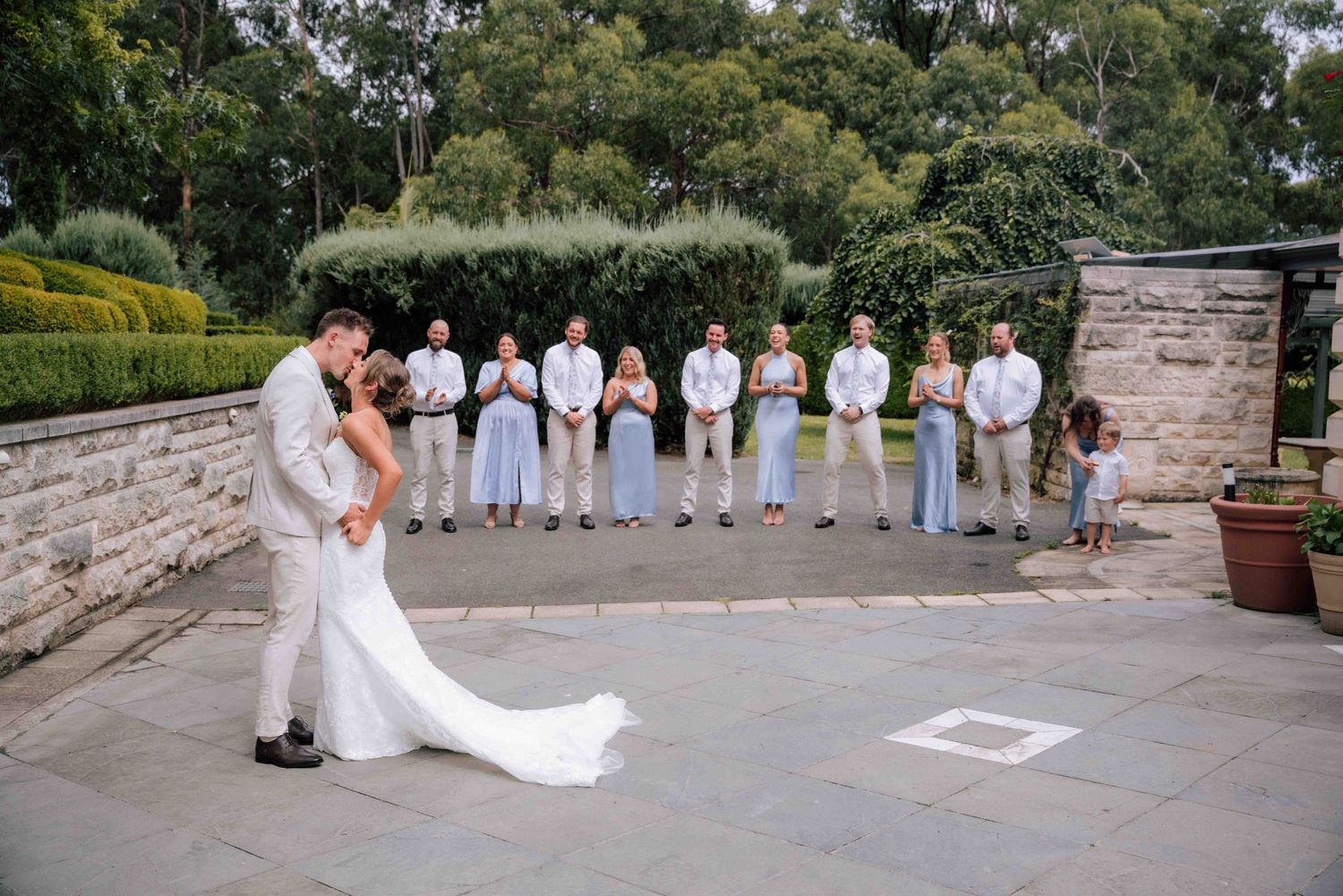 Bride and groom share first dance on stone patio while wedding party watches at outdoor garden ceremony.