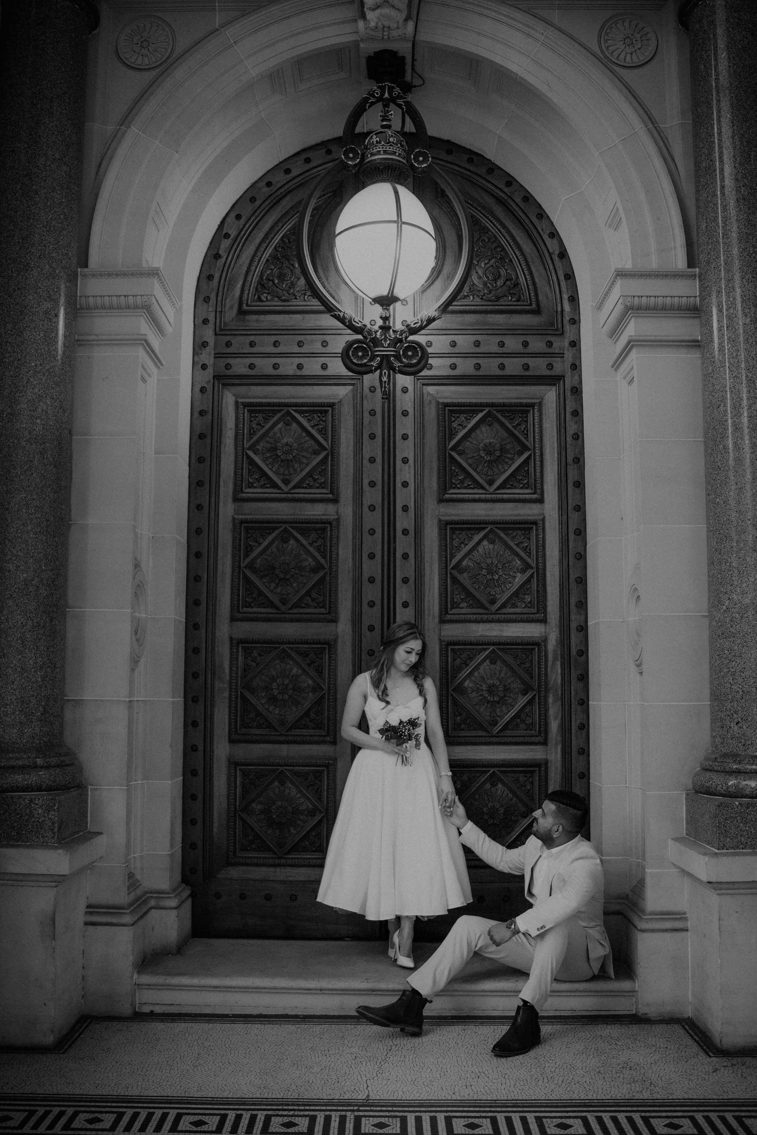 Black and white photo of bride and groom posing playfully on steps before a large decorative arched doorway.
