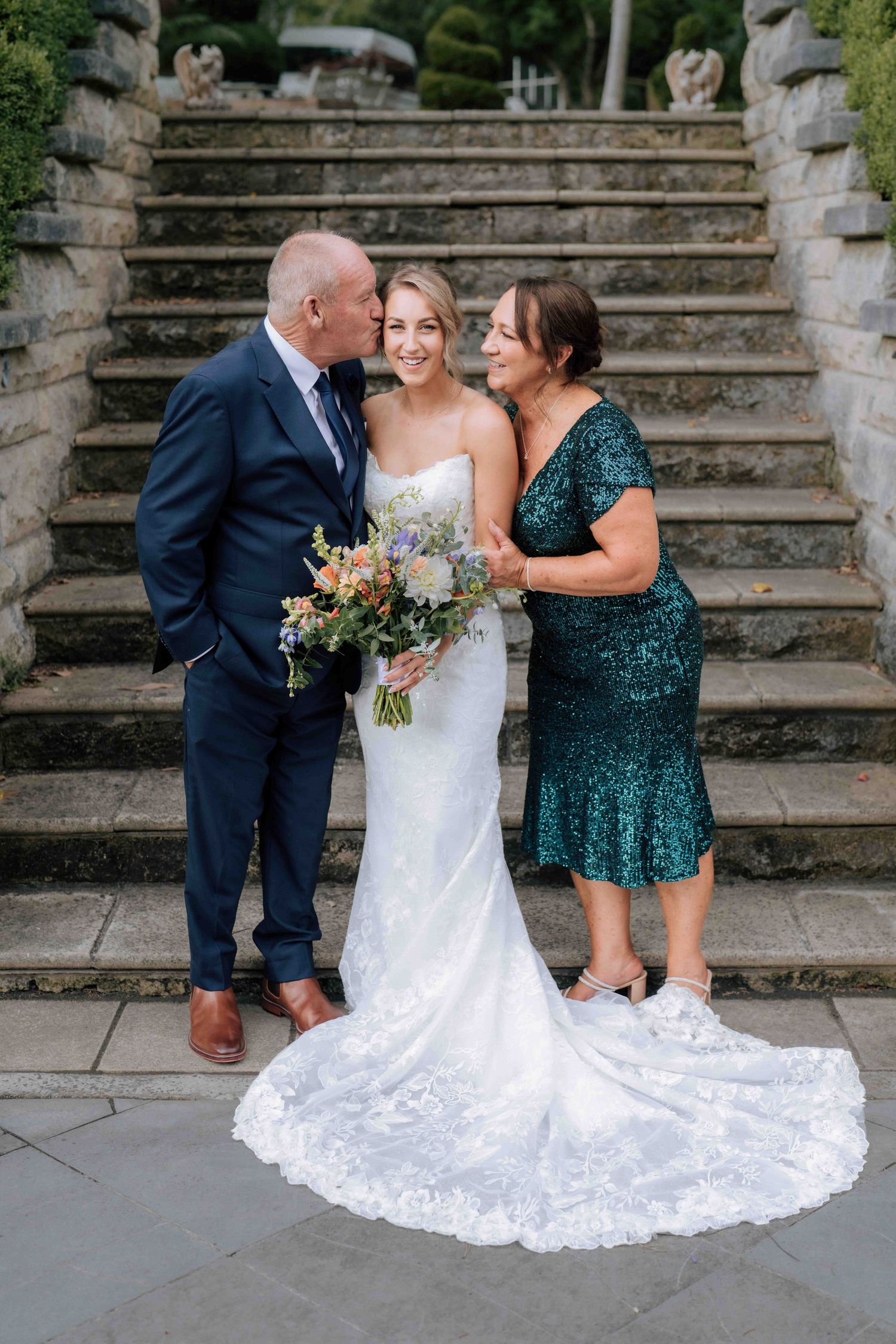 Parents lovingly kiss bride on cheeks as she holds bouquet on elegant stone staircase.