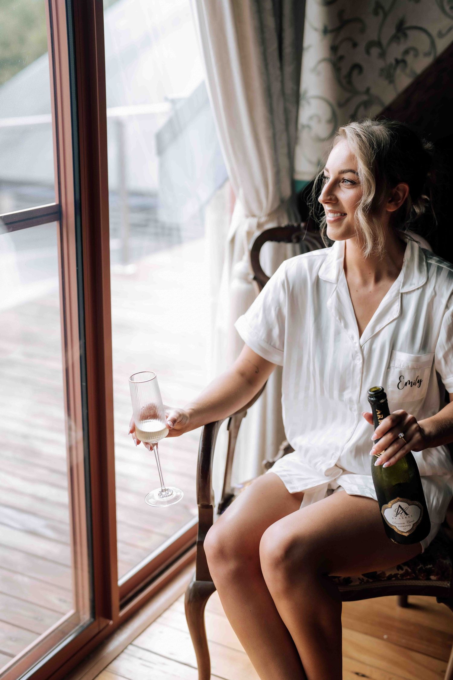 Smiling bride in white pajamas holding champagne glass and bottle by a sunlit window.