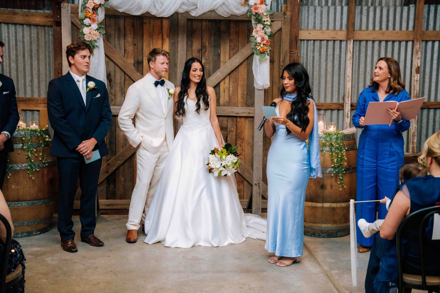 Bride and groom share a joyful moment during rustic barn wedding ceremony with wedding party and officiant.