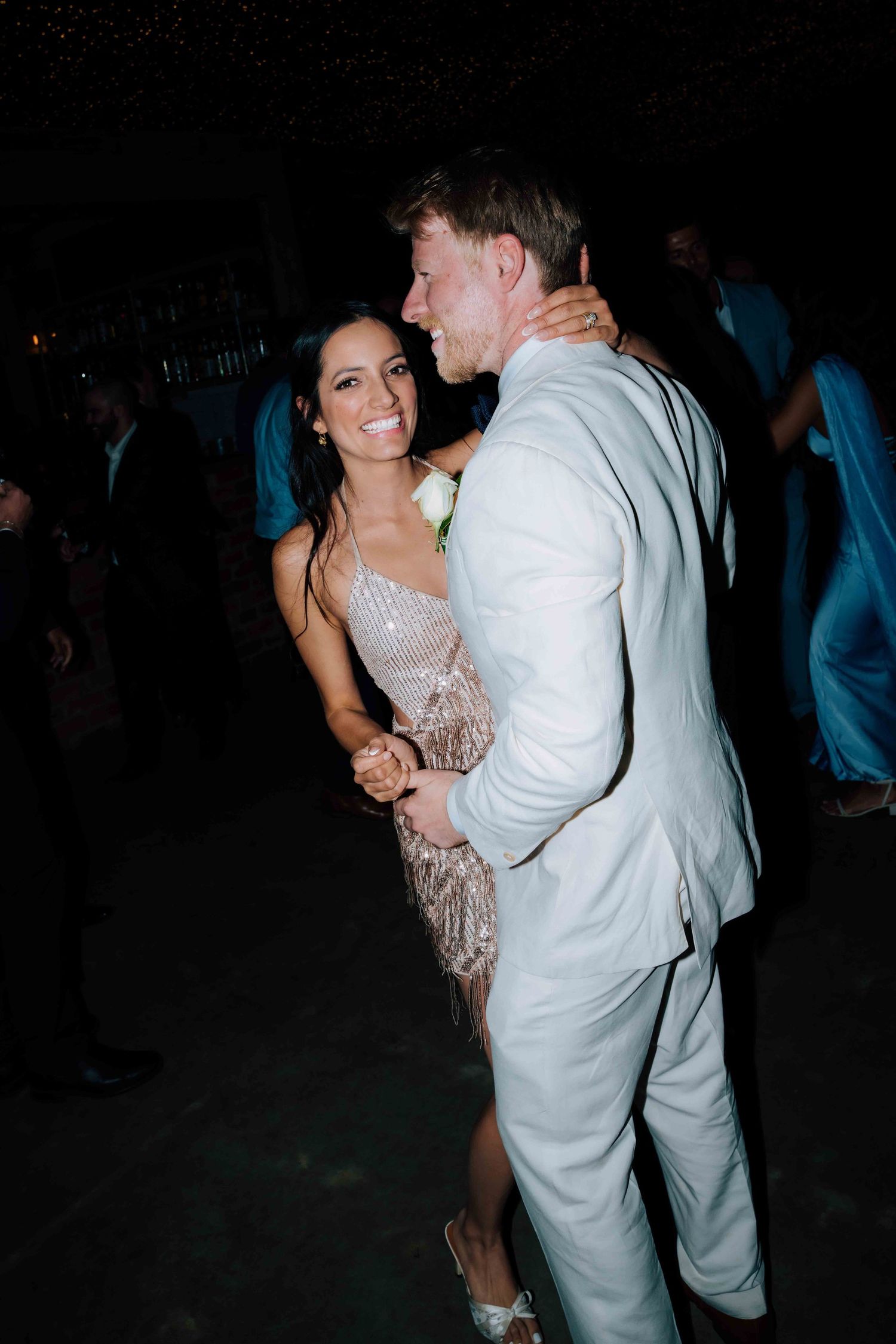 Bride and groom share a joyful first dance, she wearing a beaded fringe dress, he in a light gray suit.
