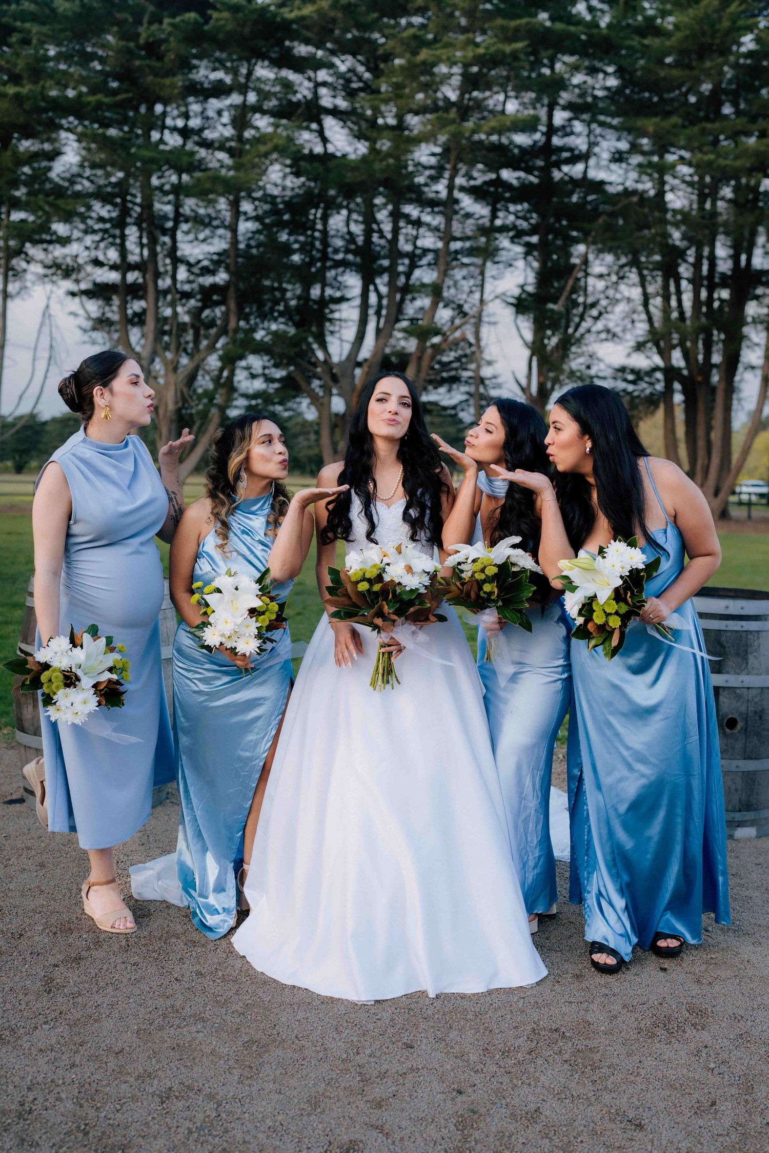 Bride in white gown poses with four bridesmaids in blue satin dresses holding bouquets outdoors.