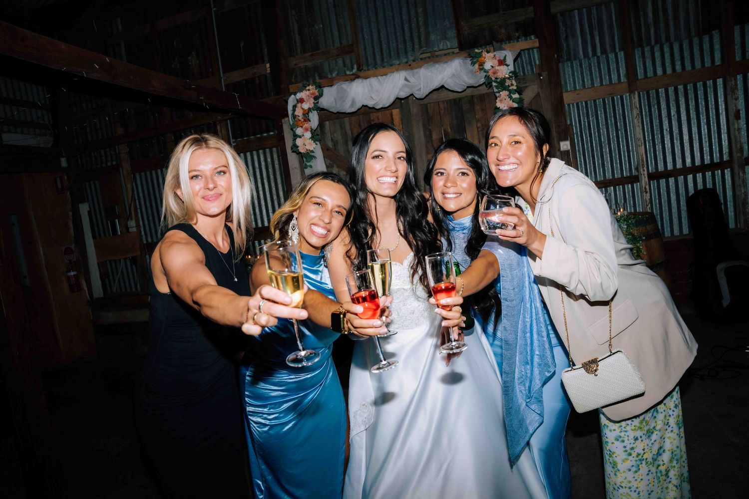 Bride in white gown celebrates with four bridesmaids holding drinks at rustic barn wedding reception.
