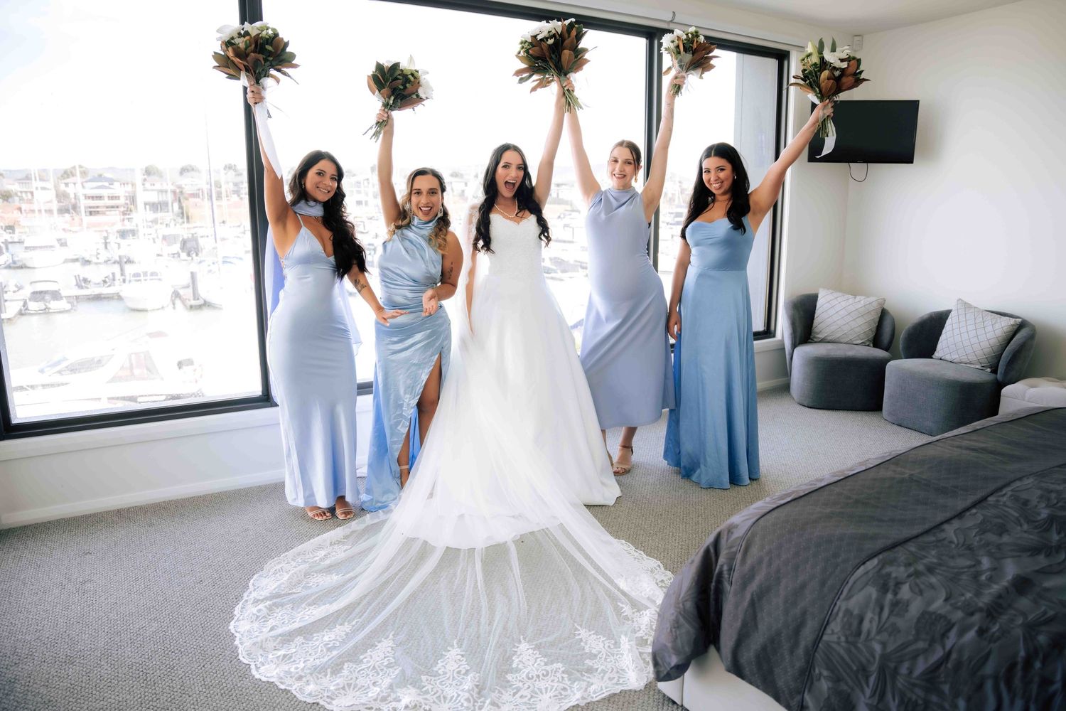 Bride in white gown and four bridesmaids in blue dresses celebrate with bouquets raised in a hotel room.