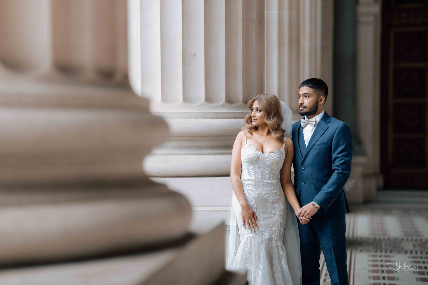 Bride and groom stand together between grand stone columns in wedding attire.