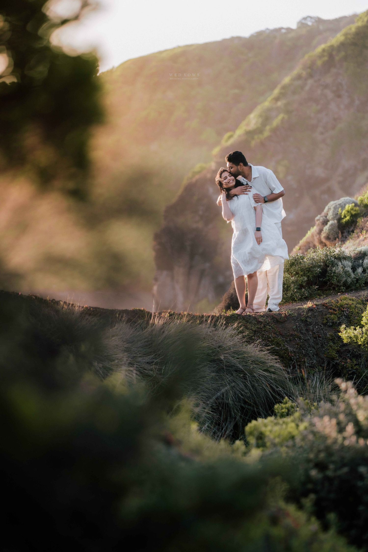 Couple in white embracing on rocky hillside surrounded by coastal vegetation at warm sunset.