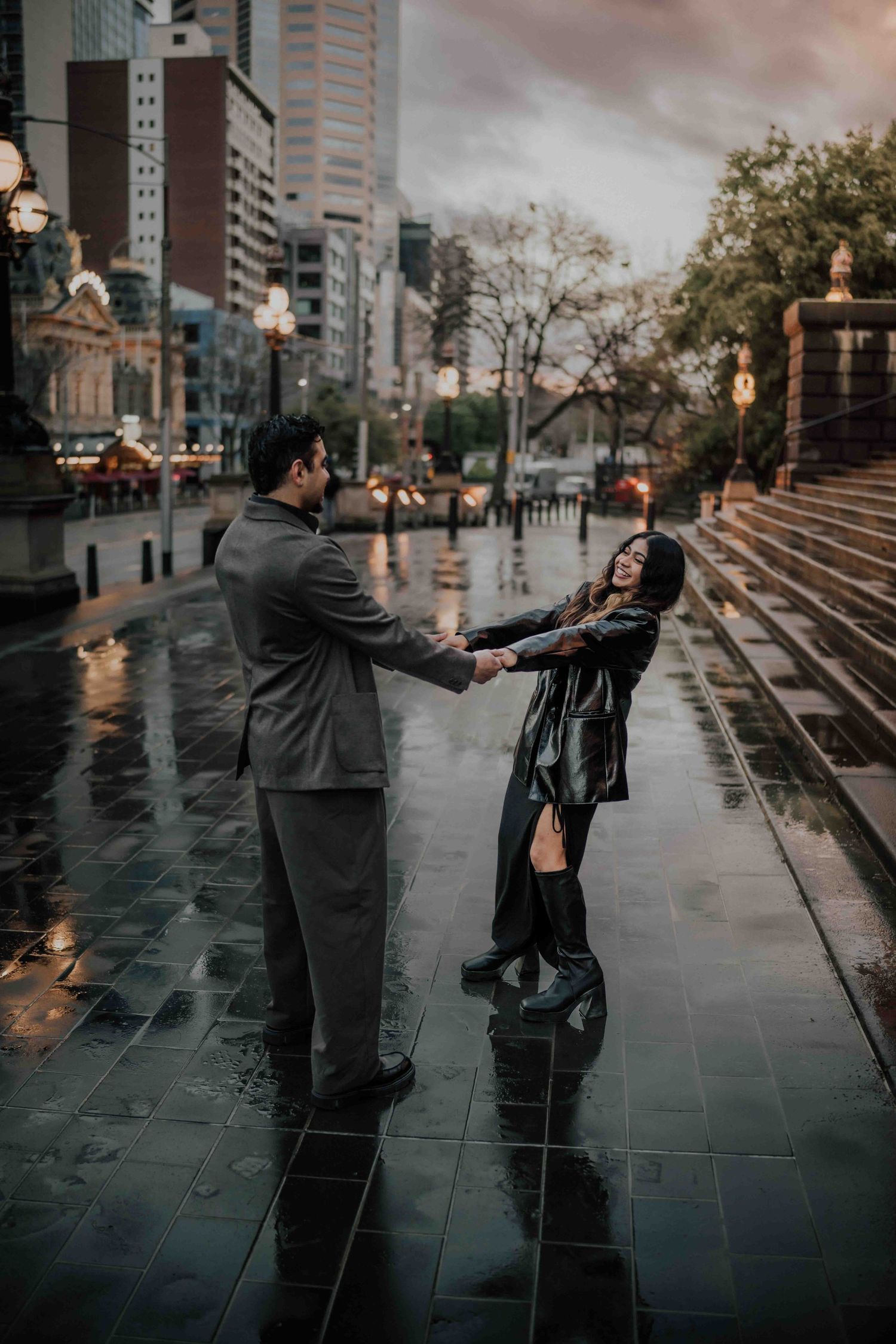 Couple laughing and holding hands on a rain-soaked city street with urban skyline backdrop.