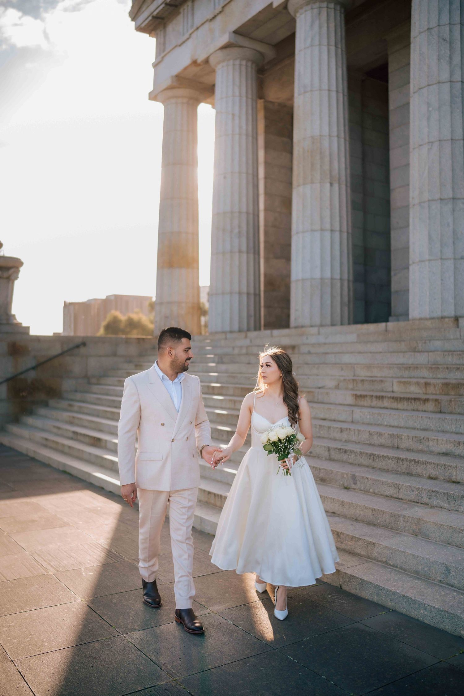 Wedding couple walks hand-in-hand beside tall stone columns in golden sunset light during bridal portraits.