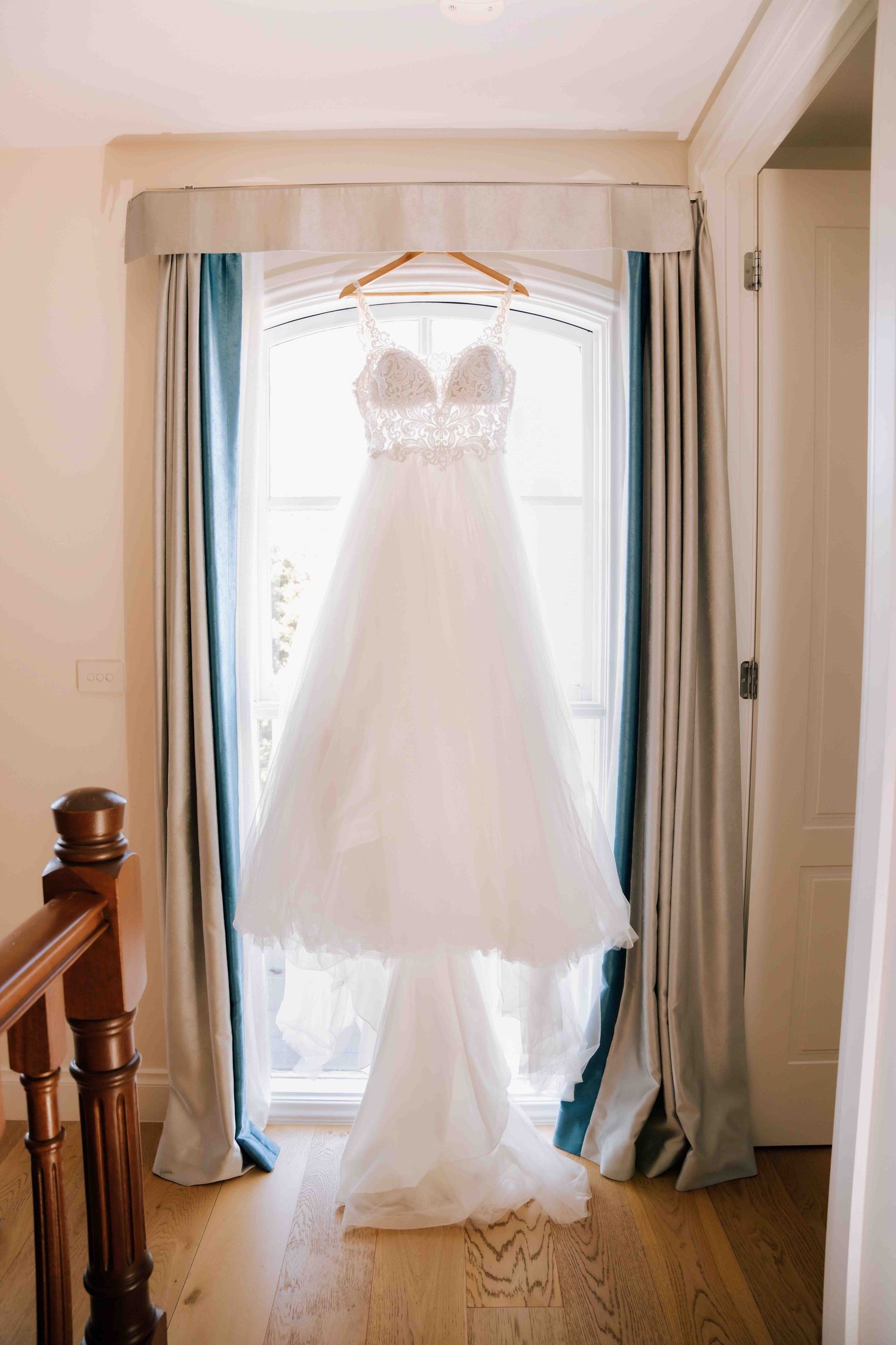 A white lace and tulle wedding dress hanging on a wooden hanger in a sunlit window.