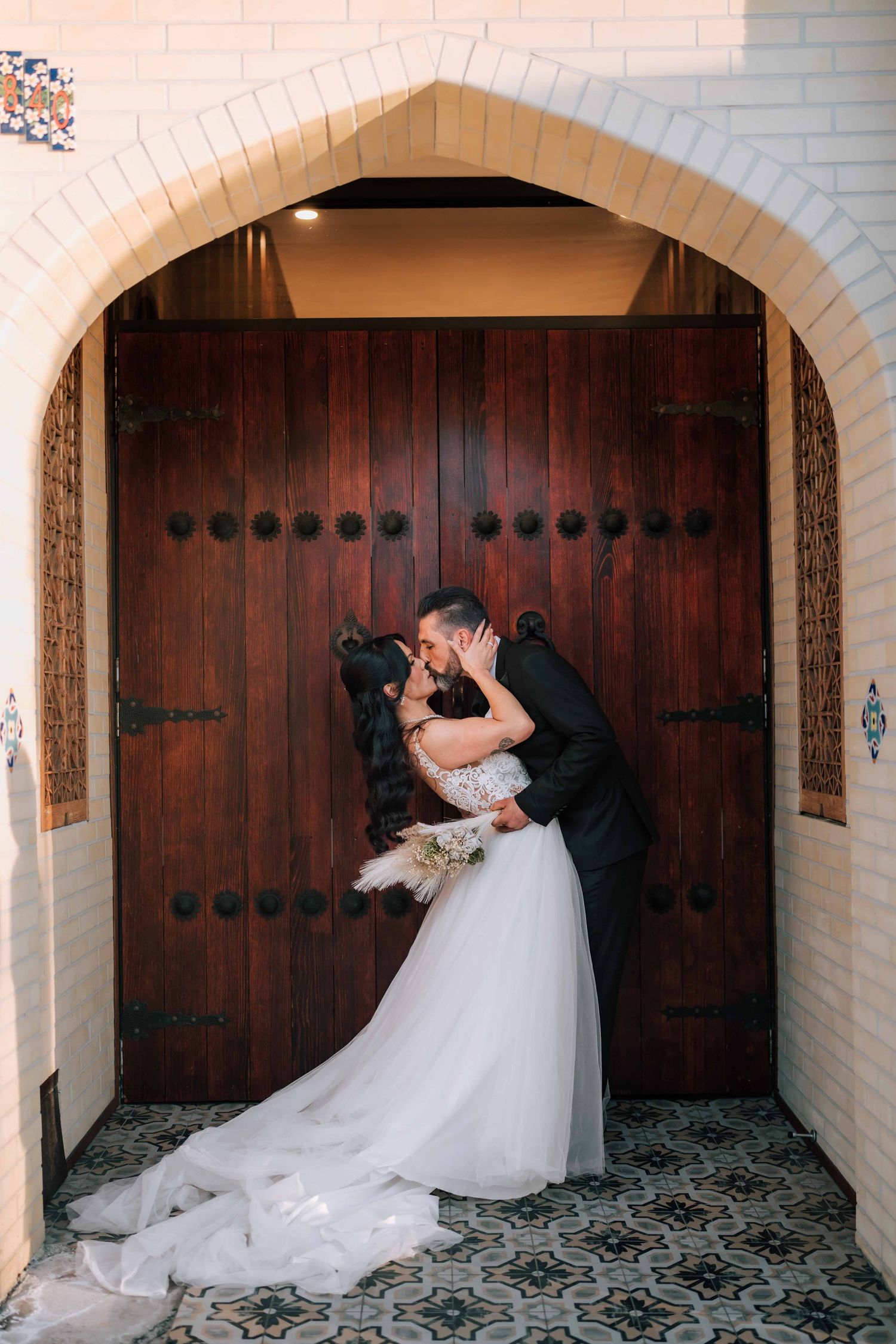 Bride and groom kissing romantically under a brick archway in front of ornate wooden doors.