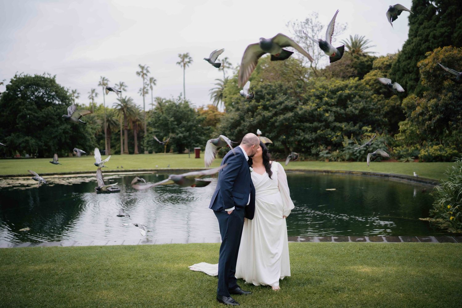 Bride and groom kissing beside a pond with birds in flight in a lush garden.