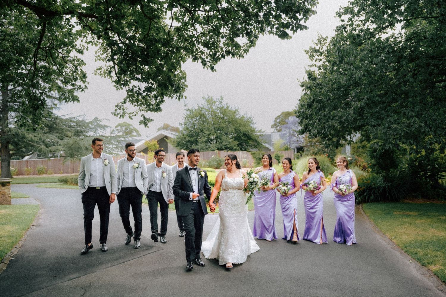 Wedding party walking in park, bridesmaids in lavender gowns, groomsmen in grey suits.