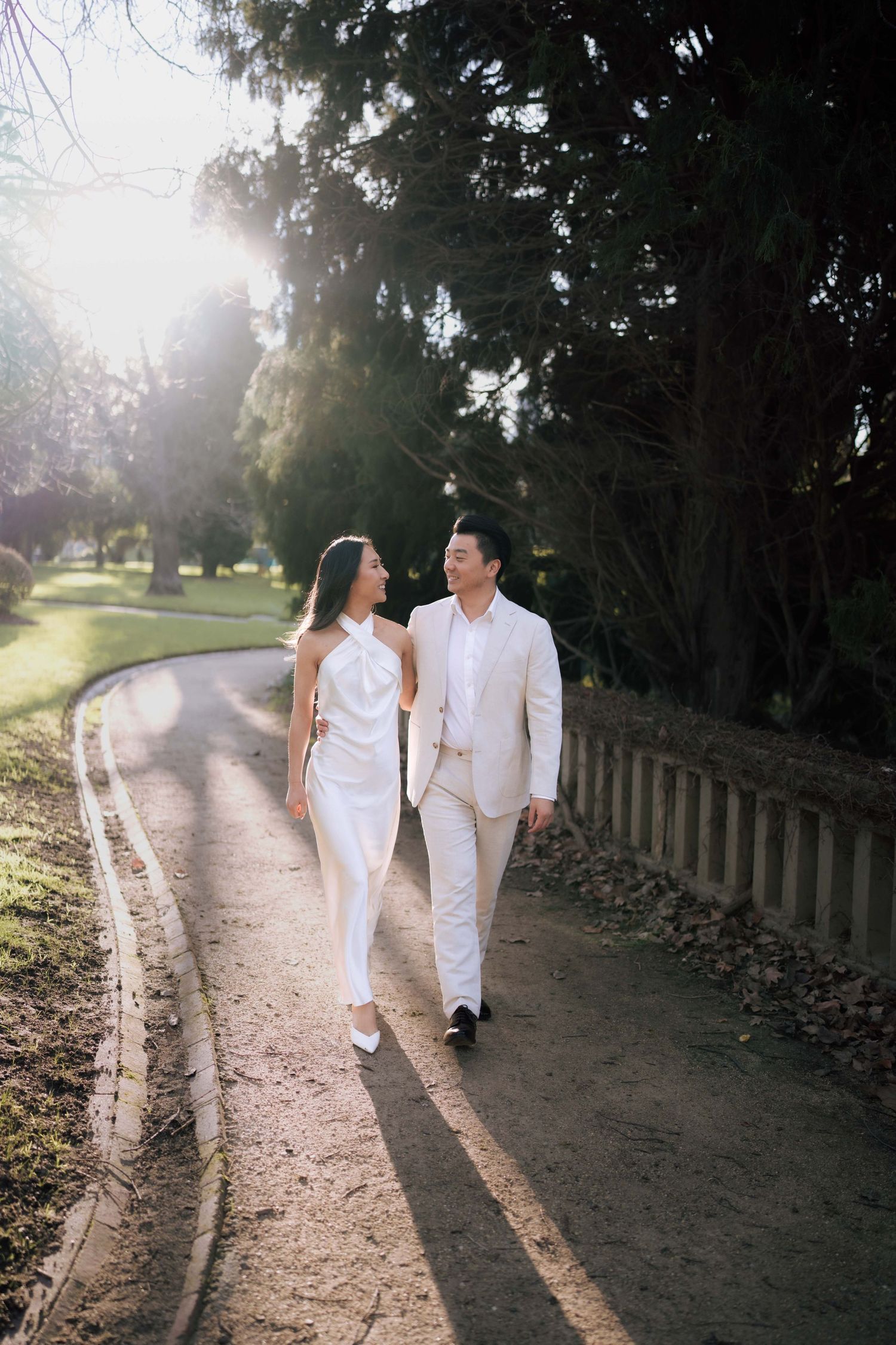 Couple in white outfits walking hand in hand along a sunlit park path surrounded by tall trees.