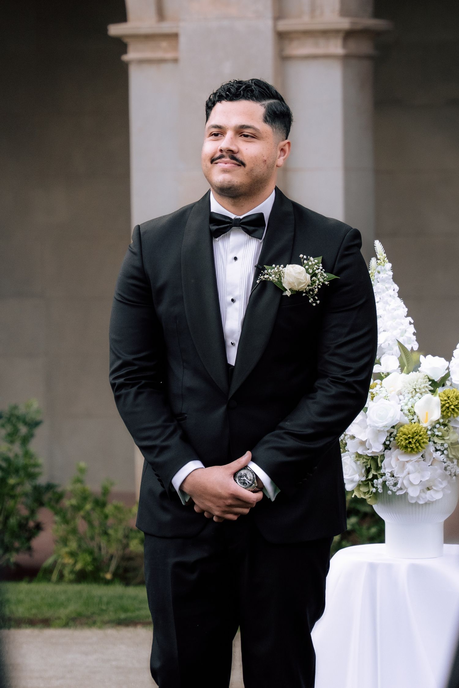 Smiling groom in black tuxedo with white rose boutonniere standing at outdoor wedding ceremony altar.