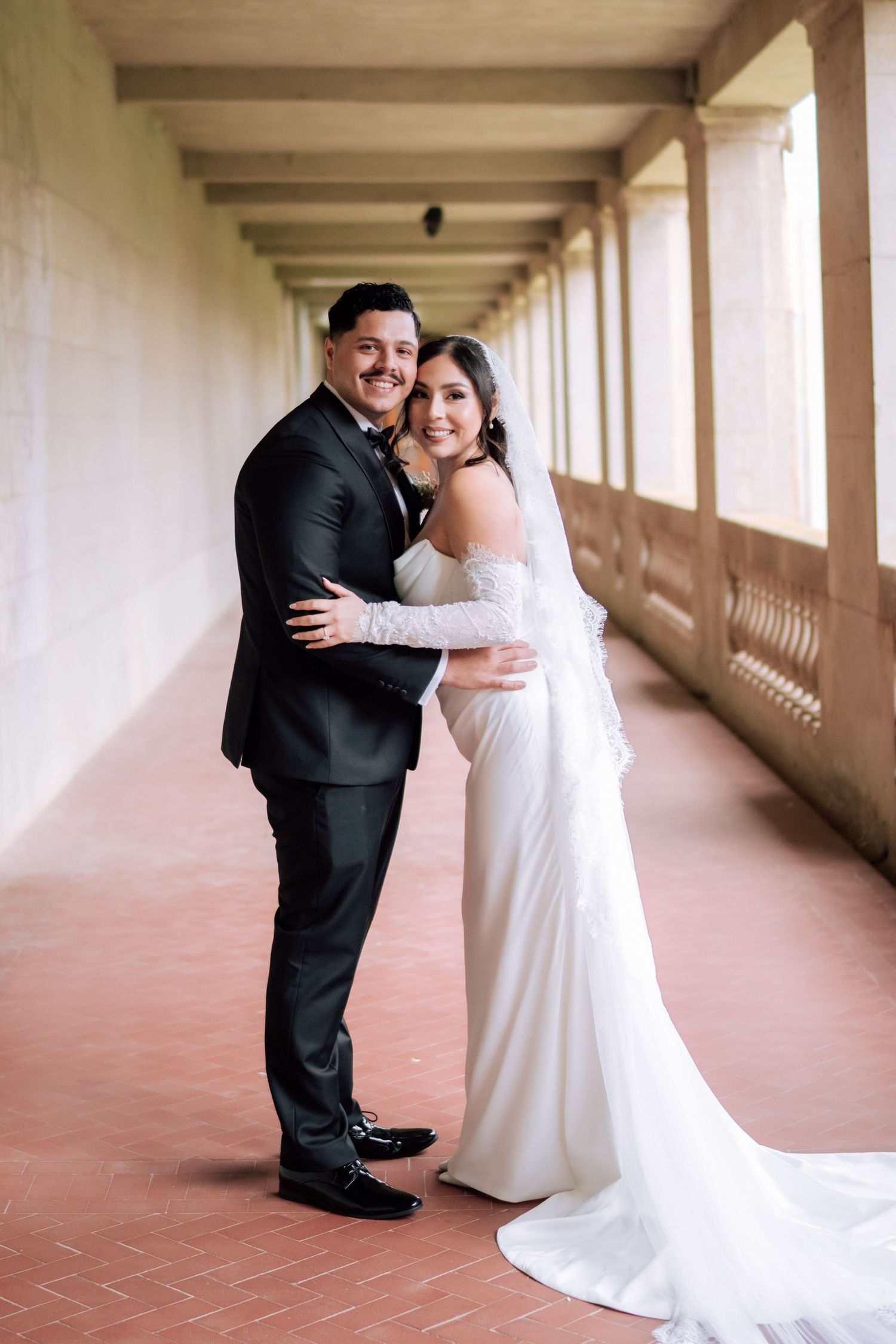 Bride in white gown and groom in black suit embracing in elegant columned hallway.