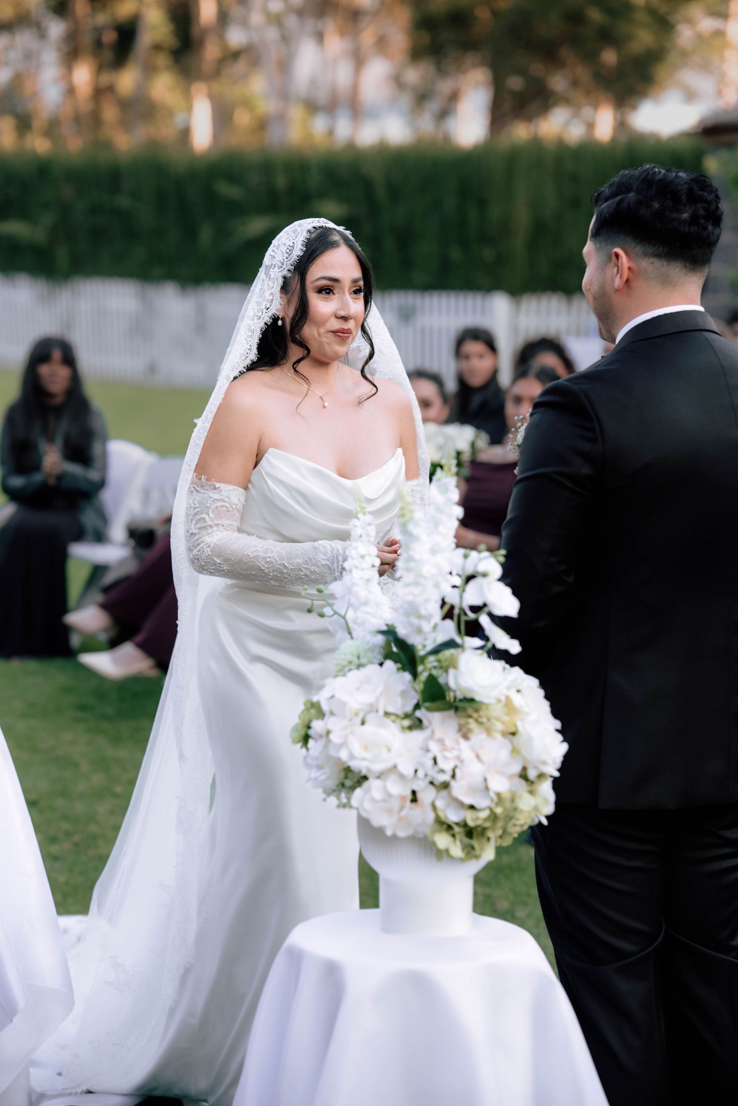 Bride in white strapless gown and lace veil stands at outdoor wedding ceremony altar with white floral arrangements.