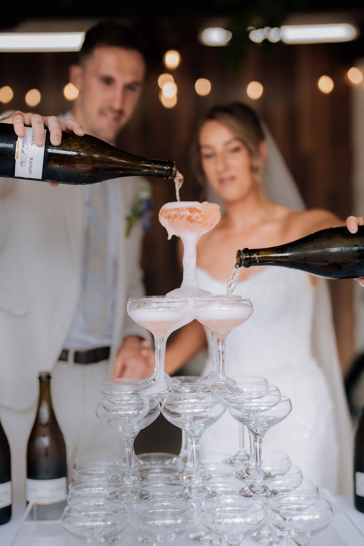 Bride and groom pouring champagne into elegant glass tower at wedding reception.